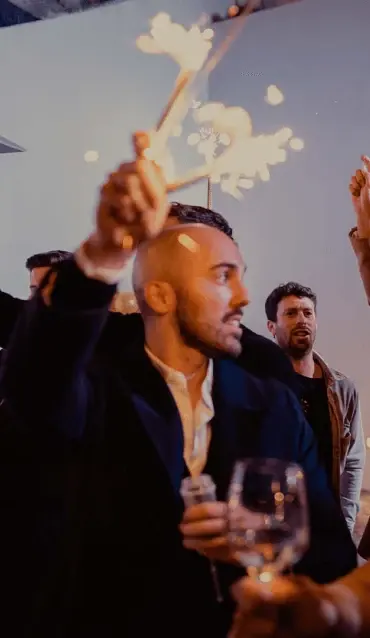 Homem de casaco escuro a levantar uma faísca de festa, com um copo na mão, durante uma celebração