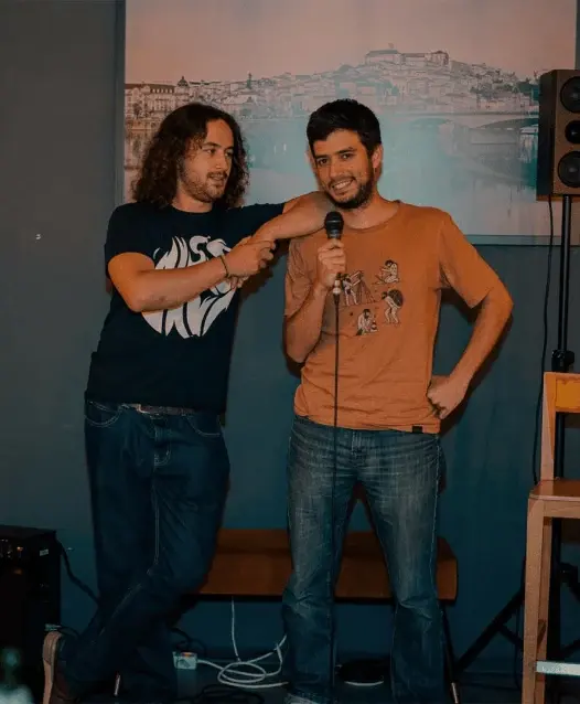 Dois humoristas em palco, um deles a segurar o microfone, em frente a um quadro com uma vista de Coimbra