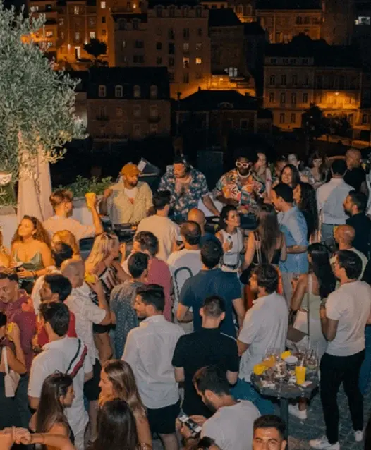 Multidão numa festa no terraço ao anoitecer, com um DJ ao fundo e casas iluminadas