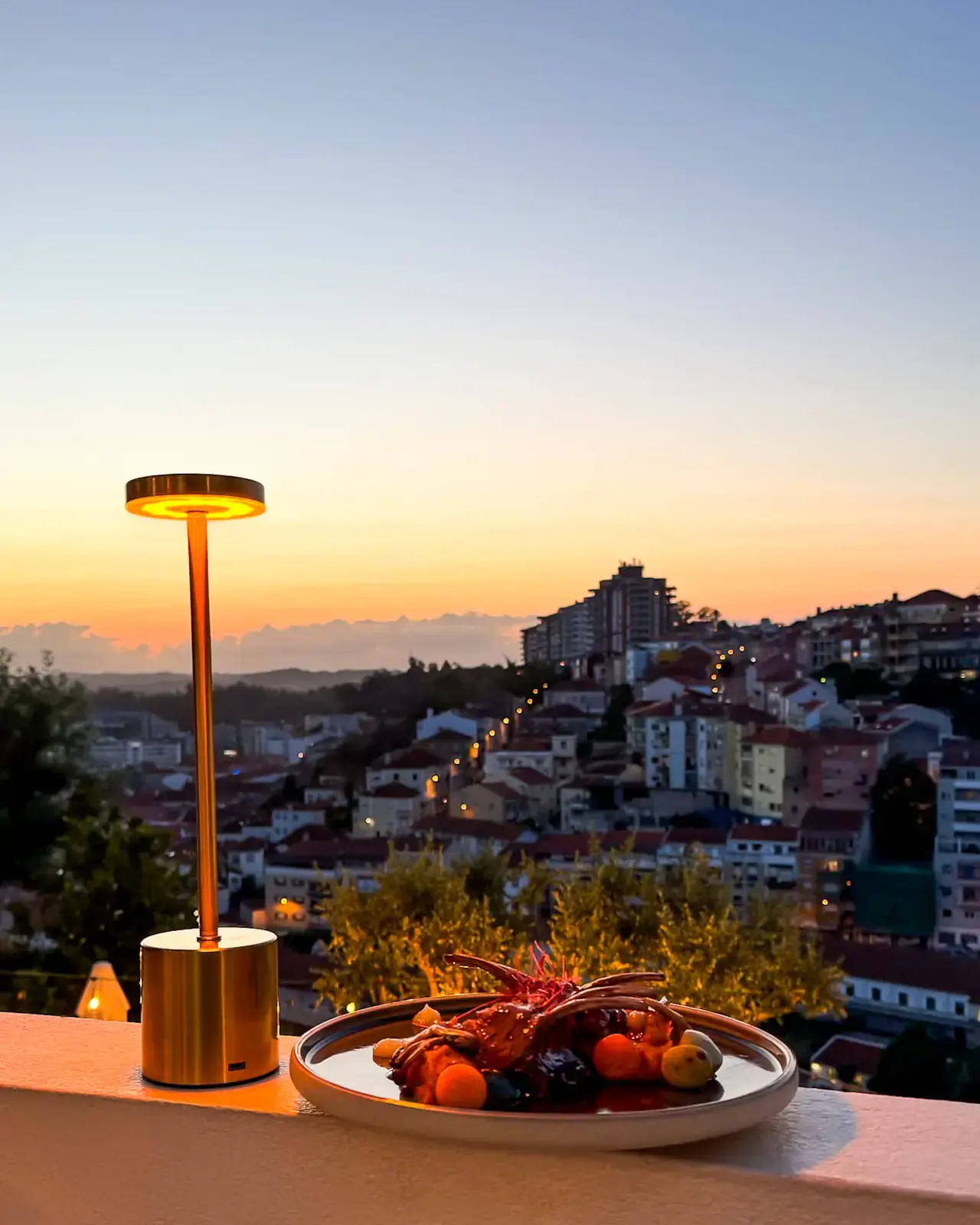Candeeiro dourado e prato de costeletas numa mureta do terraço, com Coimbra ao pôr do sol