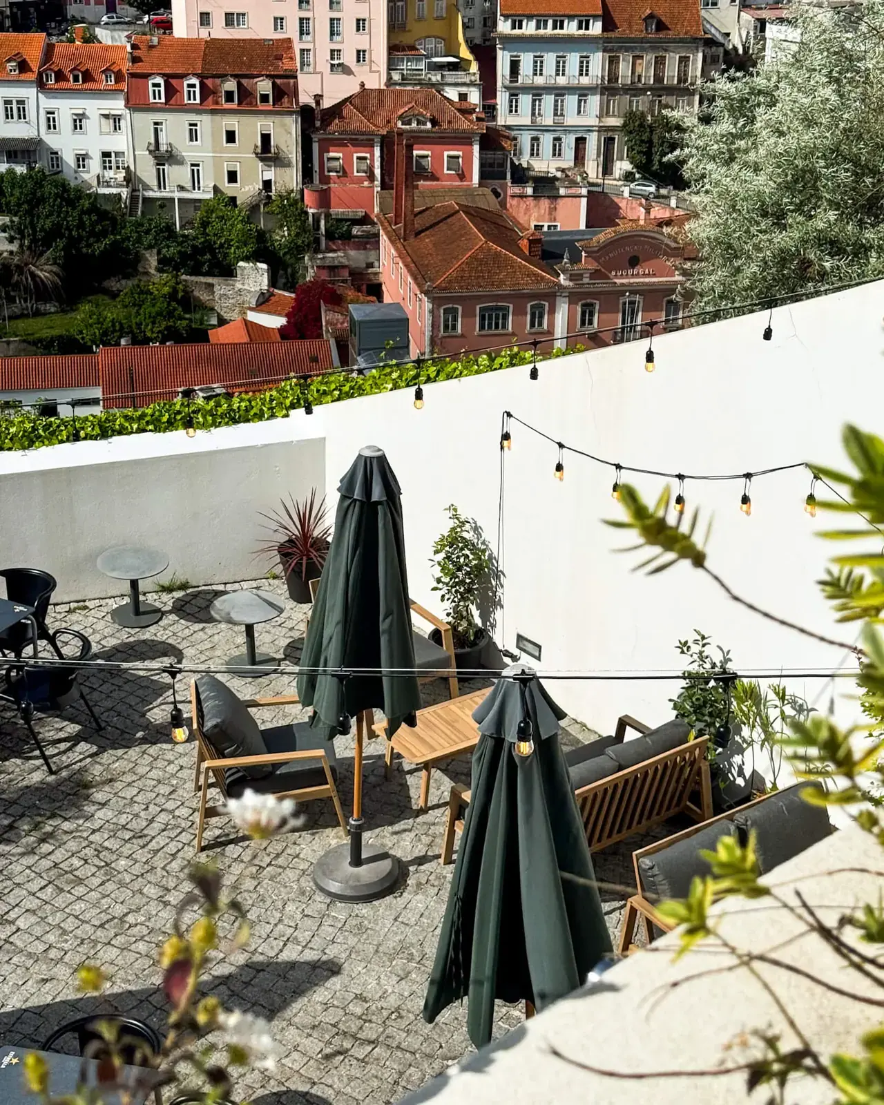 Terraço visto de cima, com mesas, sofás, chapéus de sol fechados e telhados de Coimbra ao fundo