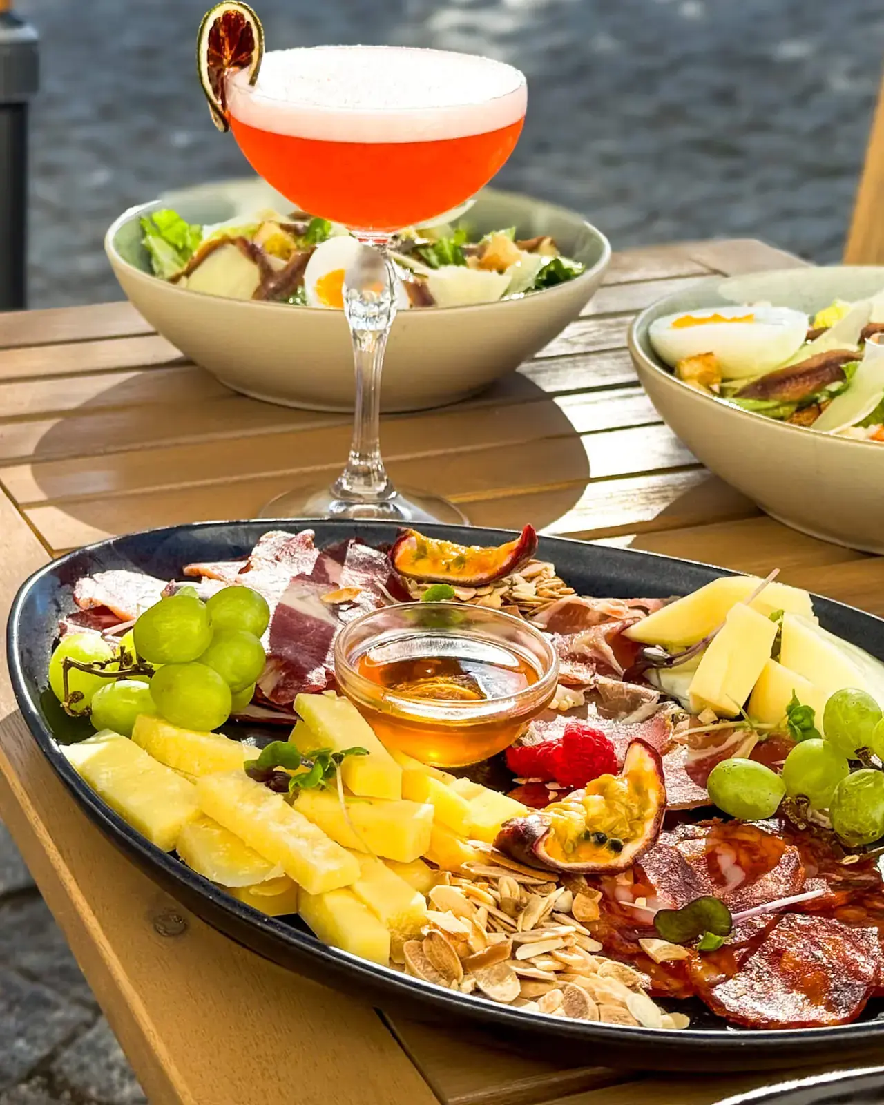 Cocktail cor de laranja com espuma ao lado de uma tábua de queijos e enchidos e de duas saladas César, numa mesa da esplanada