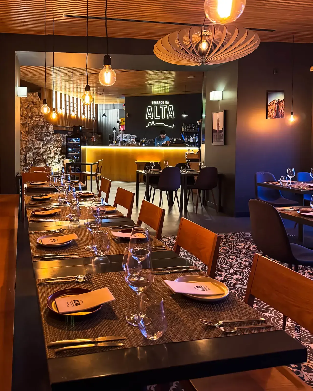 Sala interior com uma mesa comprida posta, o balcão do bar iluminado e o letreiro luminoso do Terraço da Alta