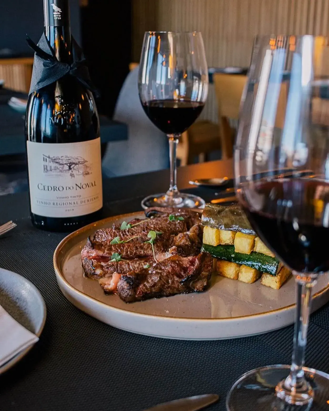 Bife fatiado com batata e courgette, uma garrafa de vinho tinto Cedro do Noval e dois copos