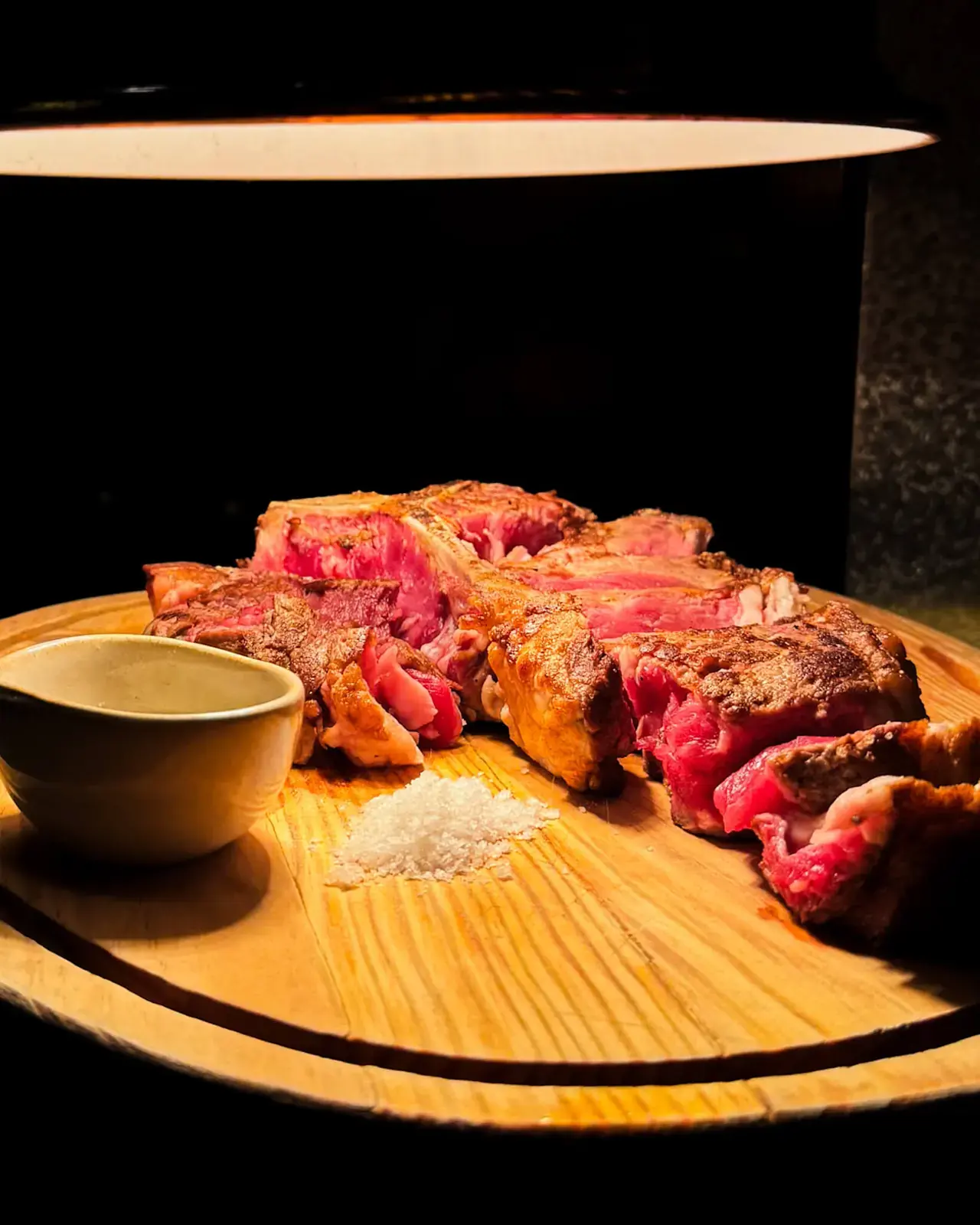 Carne grelhada fatiada numa tábua de madeira com sal grosso e uma tigela de molho, sob uma luz quente